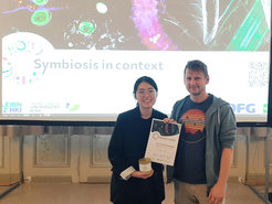 Dongik Chang mit ihrem Betreuer Tobias Engl vor einenr Präsentation mit dem Namen "Symbiosis in context". Sie hält die Preisurkund und ein Glas Honig in der Hand.